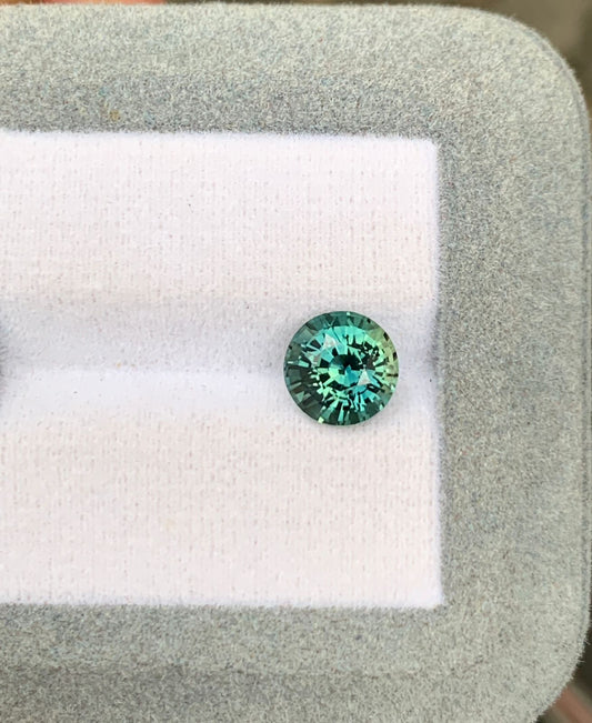 Une pierre précieuse Saphir Teal étincelante de 2,52 ct de [NOUVEAU] Bonnot Paris de Madagascar repose sur une plate-forme coussinée blanche et grise, affichant ses facettes bleu-vert vibrantes avec un fond doucement flou.
