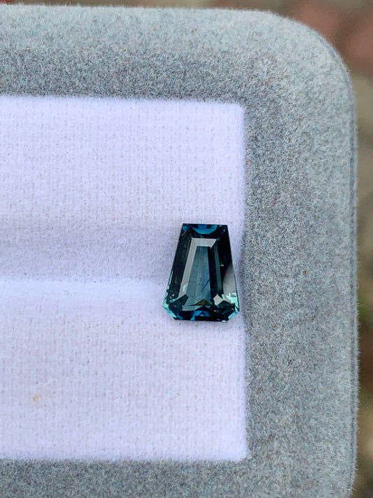 Un Saphir Teal facetté (2,52 ct) de Bonnot Paris avec une taille inhabituelle repose sur une surface blanche et coussinée dans un cadre rectangulaire gris, réfléchissant la lumière pour mettre en valeur sa clarté et sa couleur avec une élégance exceptionnelle.
