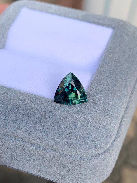 Le Saphir Teal • 2,52ct de Bonnot Paris est une pierre précieuse sarcelle triangulaire présentée sur un support coussiné gris avec un évidement, réfléchissant la lumière pour mettre en valeur les nuances de bleu et de vert.