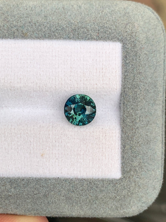 Un saphir rond sarcelle de 2,53 ct de Bonnot Paris est serti dans un coffret à bijoux gris clair doublé de velours, présentant des facettes complexes qui reflètent la lumière pour un éclat époustouflant.