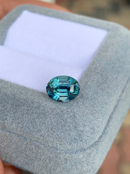 Le Saphir Teal de 2,55 carats de Bonnot Paris, une pierre bleue polie de forme ovale, est présenté dans un écrin texturé gris clair à l'intérieur blanc. Ses facettes captent magnifiquement la lumière, sublimant son éclat et sa couleur.