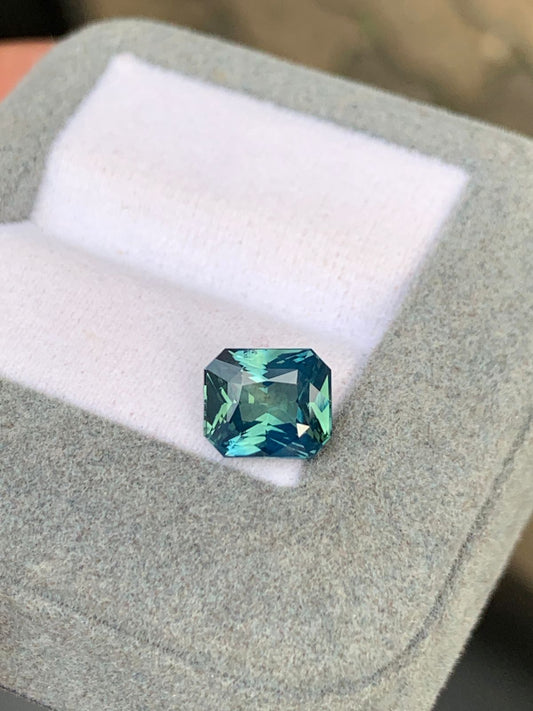 Le Saphir Teal • 3,10 carats de Bonnot Paris, une pierre précieuse rectangulaire facettée aux reflets bleus et verts, repose sur un coussin blanc dans un écrin gris ouvert. Pesant 3,10 carats, il reflète magnifiquement la lumière, soulignant sa clarté et son éclat.