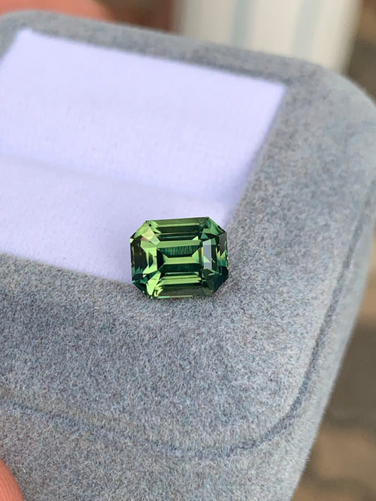 Le Saphir Teal • 3,52 ct de Bonnot Paris, une pierre précieuse verte taille émeraude, repose élégamment sur un écrin à bijoux gris clair et doux sur un fond flou.