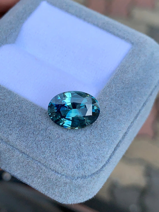 Le Saphir Teal • 6,04 carats de Bonnot Paris, une pierre précieuse bleue taille ovale étincelante, repose sur un écrin gris clair. Ses facettes reflètent brillamment la lumière, mettant en valeur différentes nuances de bleu.