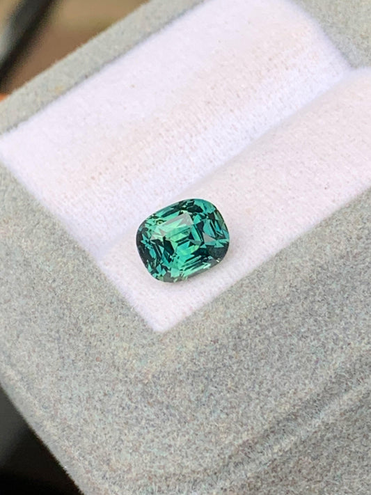 Le Saphir Teal • 2,53 carats de Bonnot Paris brille par sa taille coussin dans un écrin blanc et gris. Cette pierre rare reflète magnifiquement la lumière, mettant en valeur sa couleur vibrante et ses facettes évoquant la beauté exotique de Madagascar.