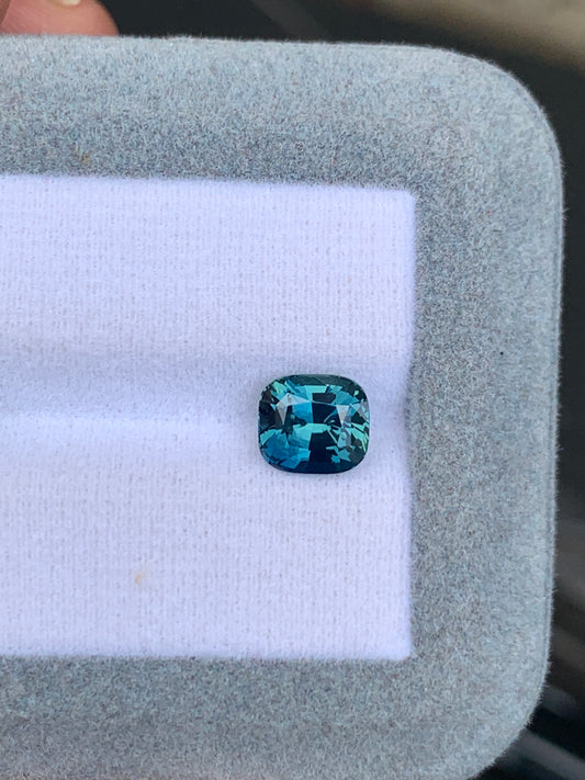 Un superbe Saphir Teal de 2,50 carats de Bonnot Paris de Madagascar, avec une taille coussin rectangulaire, repose sur un fond blanc à l'intérieur d'une boîte doublée de tissu gris, ses facettes captant magnifiquement la lumière et révélant des nuances bleues envoûtantes.