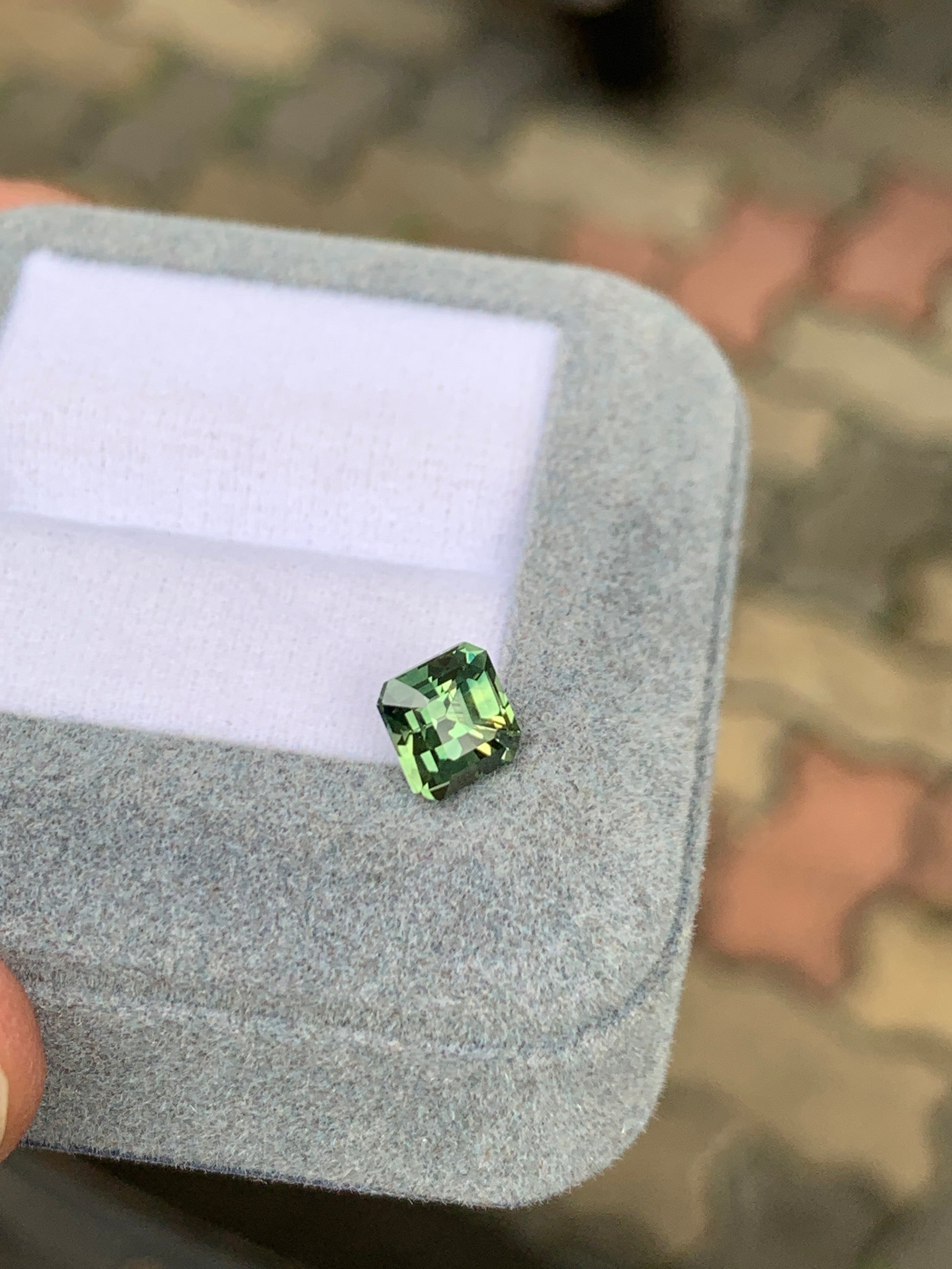 Une personne tient un Saphir Vert de 1,78 carat de [NOUVEAU] Bonnot Paris dans une boîte grise doublée de velours sur un fond de trottoir en briques flou.