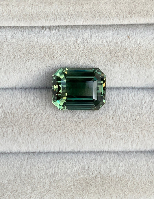 Le Saphir Vert • 6,30ct de Bonnot Paris est une pierre précieuse verte taille émeraude aux facettes complexes et à la teinte profonde et vibrante, élégamment présentée sur du velours gris.