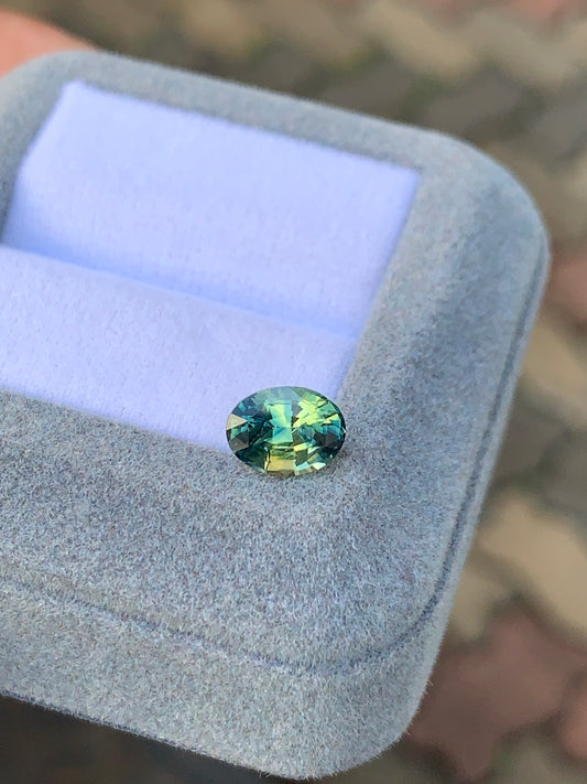 Présenté dans un écrin en peluche bleu clair, le Saphir bicolore • 2 carats de [NOUVEAU] Bonnot Paris dévoile des nuances multicolores de bleu, de vert et de jaune. Le fond flou laisse entrevoir une surface à motifs.