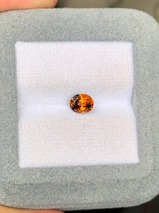 Le Saphir bicolore • 1,13 ct de Bonnot Paris, une pierre précieuse ronde et étincelante d'un orange vif rappelant une pierre précieuse du Sri Lanka, est présenté sur un coussin blanc moelleux dans un cadre gris. Sa teinte vibrante reflète magnifiquement la lumière sur le coussin lisse.