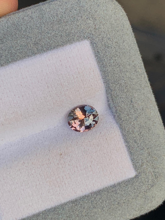 Un saphir bicolore rond et brillant de 1,20 ct du Sri Lanka, aux teintes rosées et aux reflets arc-en-ciel, repose sur un tissu blanc rembourré de mousse grise douce de [NOUVEAU] Bonnot Paris.