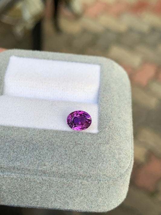 Une pierre précieuse ovale violette étincelante, semblable au Saphir bicolore • 1,52 ct de Bonnot Paris, repose élégamment sur un coussin blanc dans un écrin gris. La surface floue et colorée de l'arrière-plan met en valeur cette exposition de pierres précieuses.