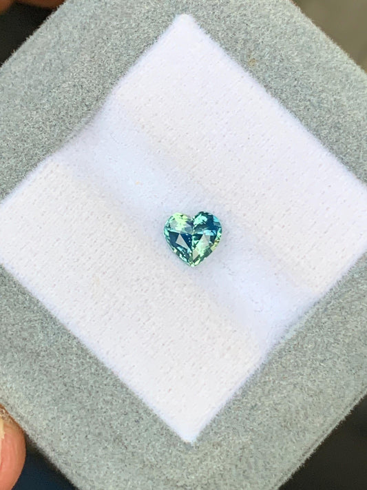 Un saphir bicolore en forme de cœur de 1,06 ct bleu et vert de Bonnot Paris, niché sur un coussin blanc à l'intérieur d'une boîte grise, provient du Sri Lanka.