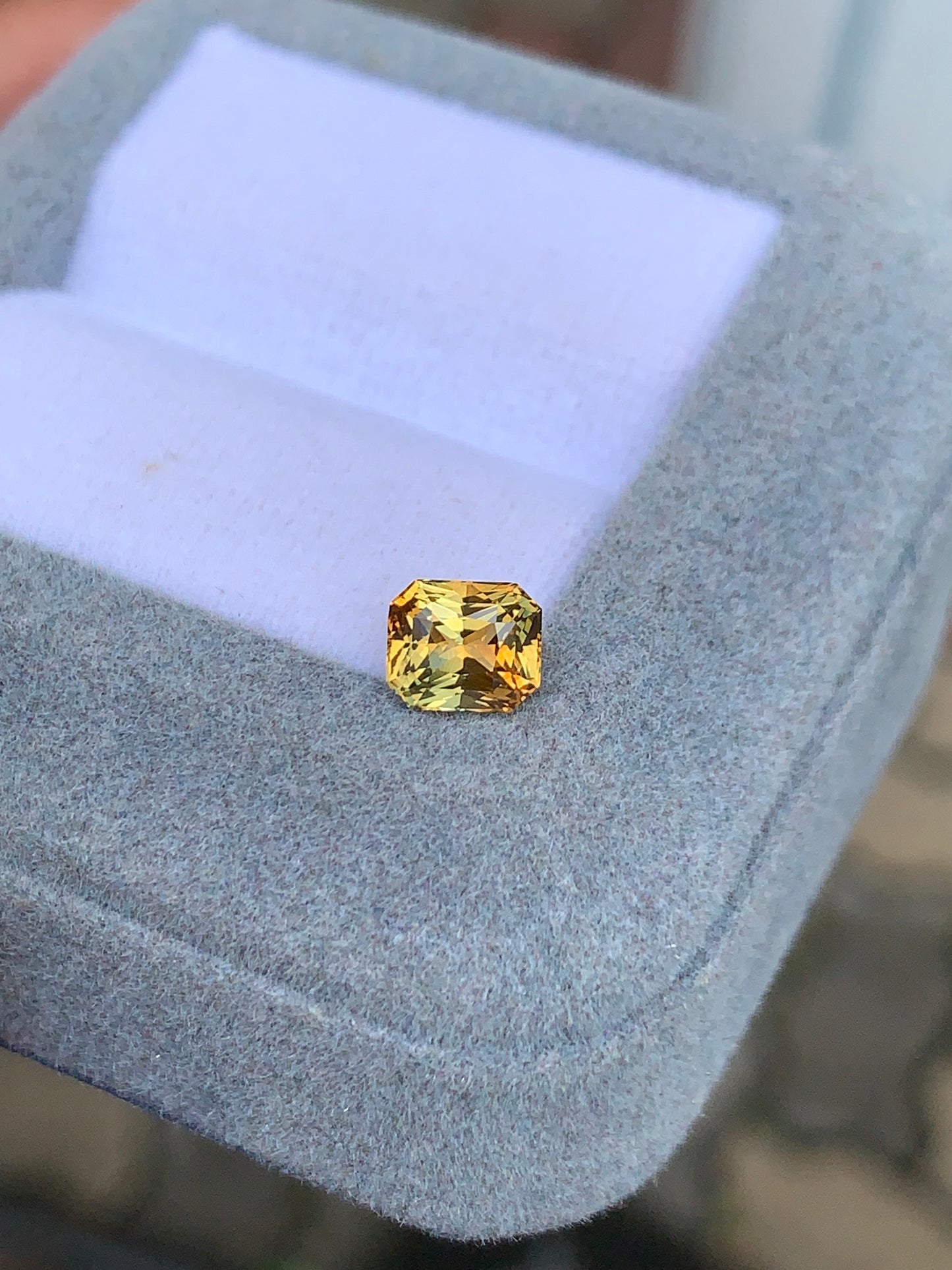 Le « Saphir bicolore • 1,10 ct » de Bonnot Paris est une magnifique pierre jaune rectangulaire aux angles arrondis, présentée avec élégance dans un écrin gris à l'intérieur en tissu blanc. Ses facettes rayonnent à la lumière naturelle, soulignant son exceptionnelle pureté.