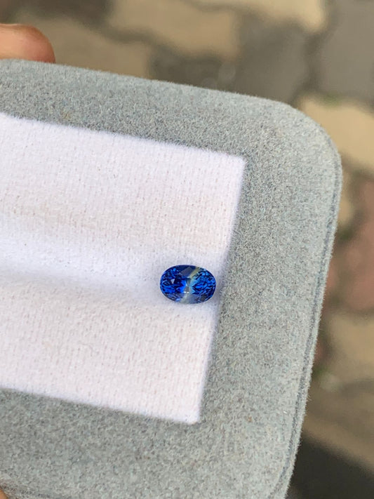Gros plan sur le Saphir bicolore 1,10 carat de Bonnot Paris. Cette pierre ovale bleue présente des motifs rappelant une carte du monde, sur un fond gris et blanc doux. Le pouce est partiellement visible.