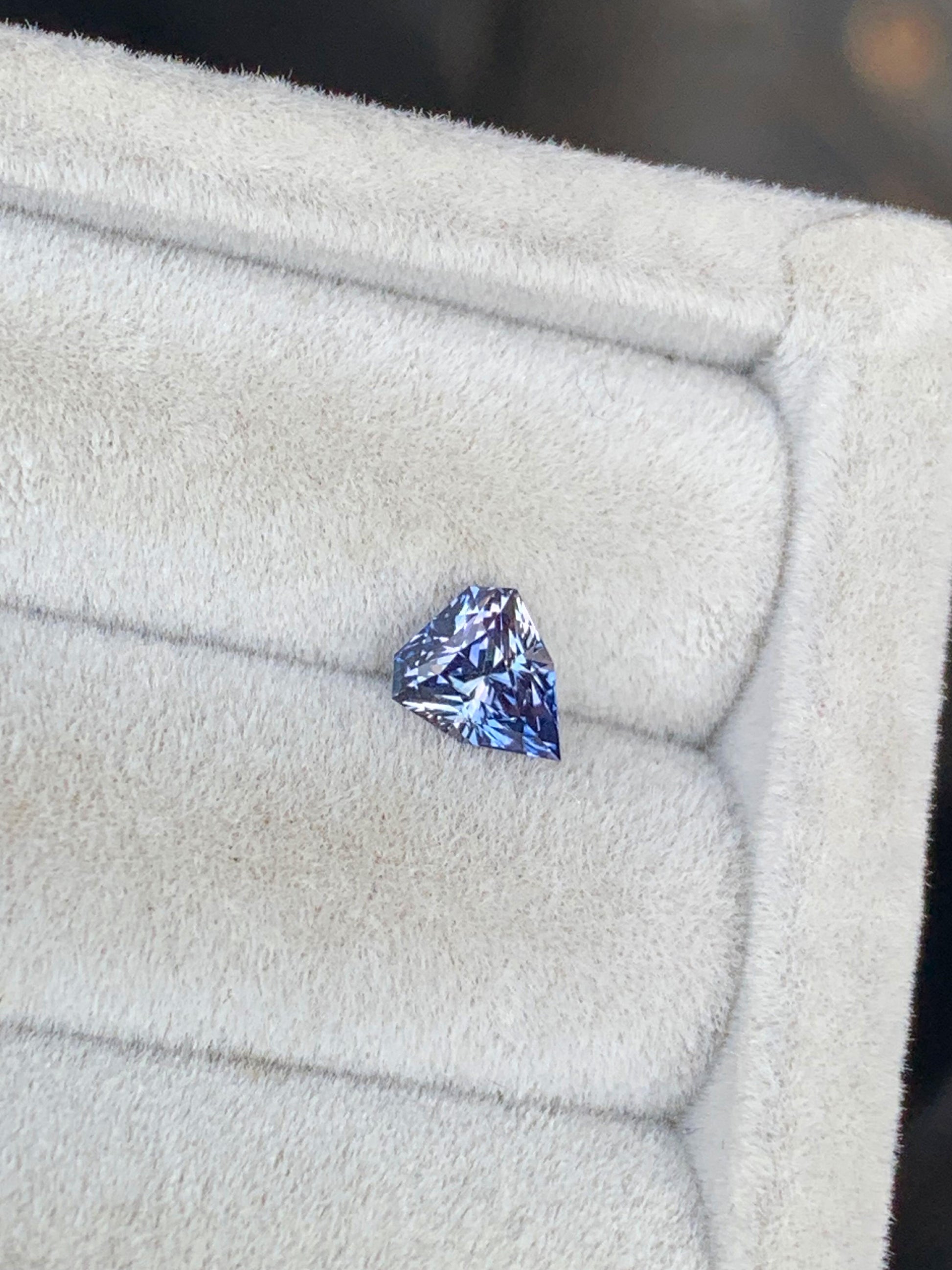 Un petit saphir bicolore triangulaire de 1,15 ct de Bonnot Paris, présentant des touches de violet, est élégamment positionné légèrement à gauche sur un plateau à bijoux texturé gris clair, illustrant la joaillerie d'exception.
