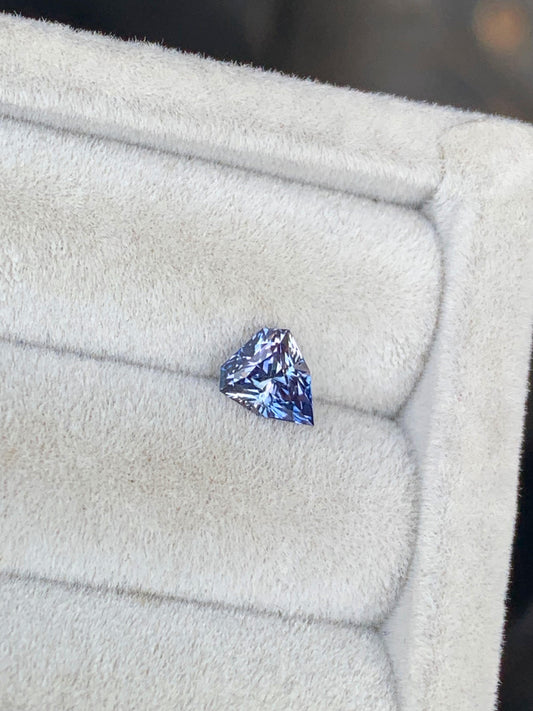 Un petit saphir bicolore triangulaire de 1,15 ct de Bonnot Paris, présentant des touches de violet, est élégamment positionné légèrement à gauche sur un plateau à bijoux texturé gris clair, illustrant la joaillerie d'exception.