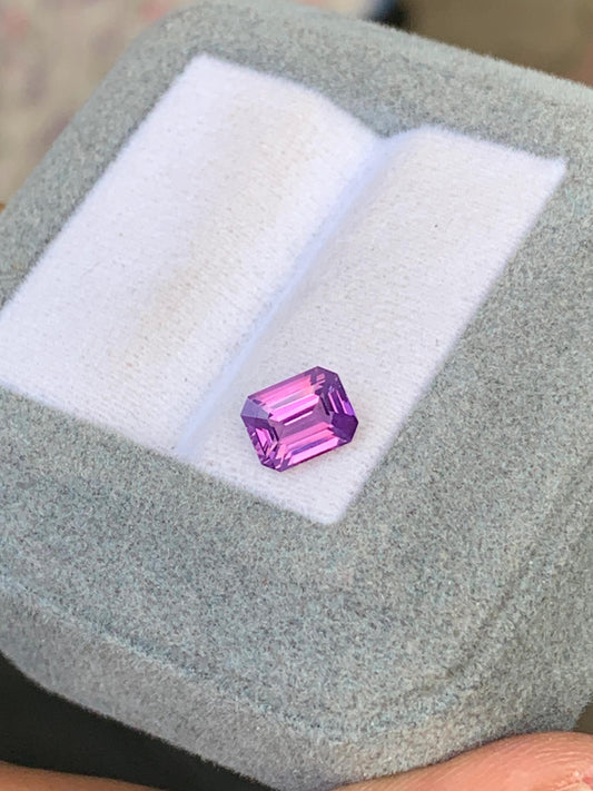 Le Saphir bicolore • 1,51 ct de [NOUVEAU] Bonnot Paris est une pierre précieuse violette taille émeraude éclatante, posée sur un coussin blanc dans un écrin gris clair. Ses facettes éclatantes et polies sont magnifiquement mises en valeur par des reflets.