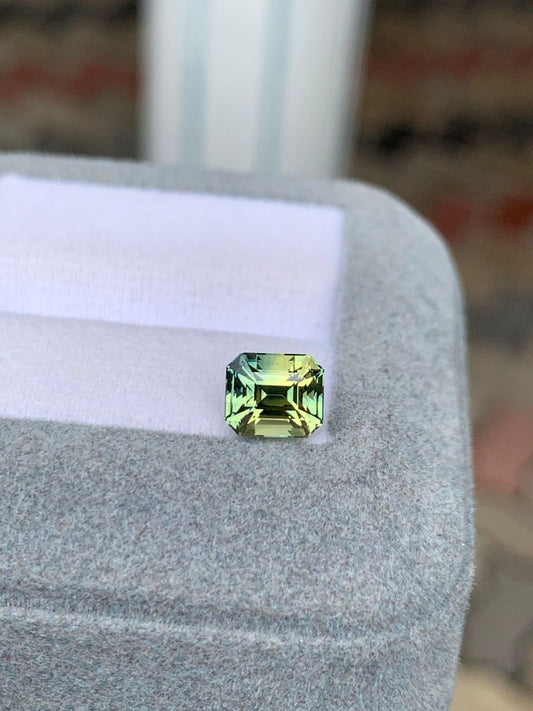 Le Saphir bicolore, une pierre précieuse de 2,00 carats de Bonnot Paris, affiche une teinte vert clair avec des nuances vertes variées alors qu'il scintille au sommet d'une boîte à bijoux grise en velours de Madagascar.