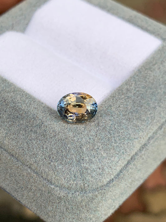 Un Saphir bicolore • 2,02ct de Bonnot Paris, aux teintes ovales de bleu, vert et marron clair, repose sur une surface grise texturée avec un fond légèrement flou.