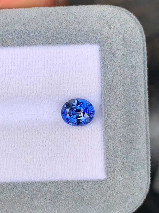 Le Saphir bicolore • 2,10ct de [NOUVEAU] Bonnot Paris, un exquis saphir bleu ovale du Sri Lanka, est magnifiquement mis en valeur sur une surface en tissu blanc et gris.