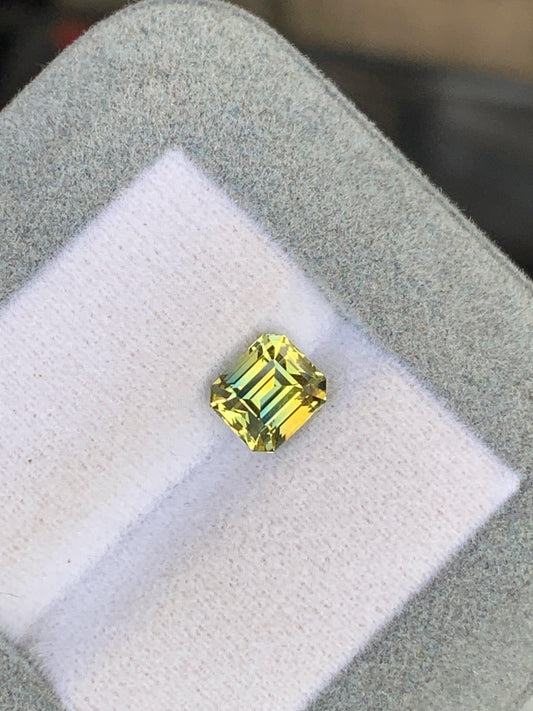 Un Saphir bicolore taille carrée • 2,18ct de [NOUVEAU] Bonnot Paris, ressemblant à des pierres précieuses rares d’exception de Madagascar, est élégamment présenté sur un tissu blanc dans une élégante boîte grise.
