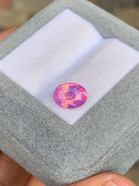 Un Saphir bicolore ovale rose vibrant • 2,50 ct de [NOUVEAU] Bonnot Paris, rappelant une pierre précieuse sri-lankaise rare, est présenté sur une boîte grise avec un intérieur blanc, tenu dans la main de quelqu'un.