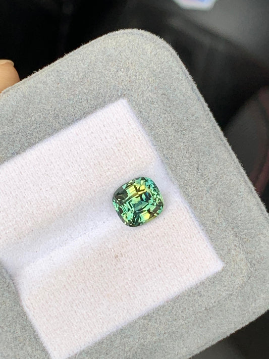 La pierre précieuse verte taille coussin de 2,58 ct avec une teinte jaune, Saphir bicolore de Bonnot Paris, est présentée dans un coffret carré en velours gris avec un intérieur blanc doux.