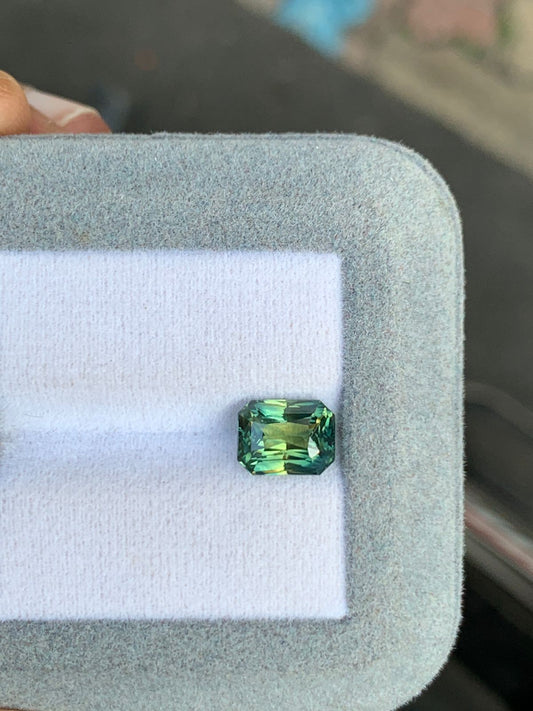 Une personne tient un coffret à bijoux gris matelassé contenant une pierre non chauffée taille émeraude aux nuances vertes et bleu sarcelle. Ce saphir bicolore de 3,02 ct est serti sur un intérieur en tissu blanc de la marque Bonnot Paris, dont l'arrière-plan flou renforce l'attrait.
