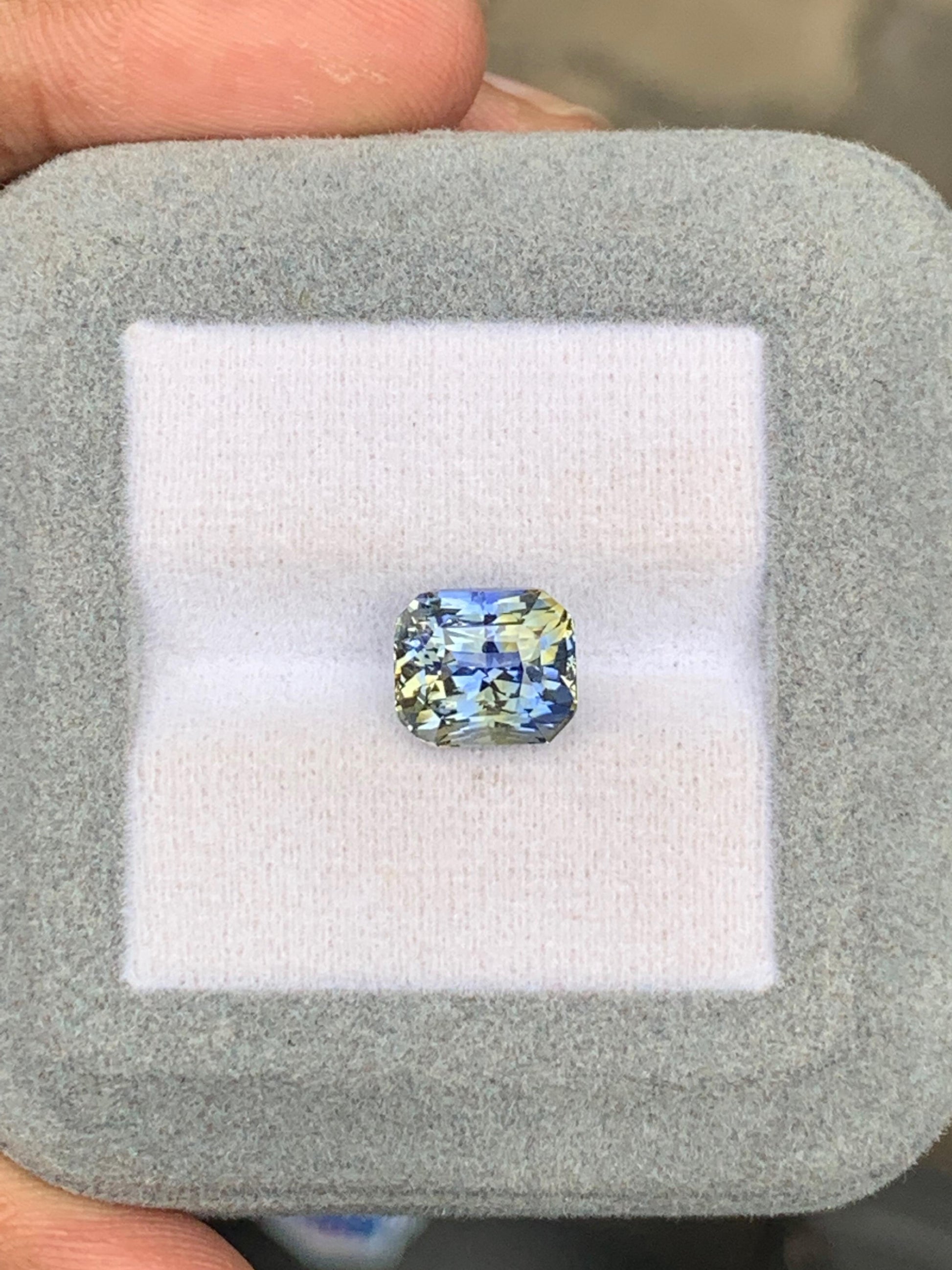 Un saphir bicolore de 5,01 carats signé Bonnot Paris, aux reflets bleu-jaune, est niché dans un écrin gris doux. Des doigts délicatement saisis mettent en valeur les facettes complexes de la pierre, évoquant les éclatantes pierres précieuses du Sri Lanka.