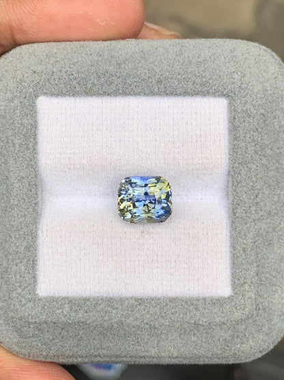 Un saphir bicolore de 5,01 carats signé Bonnot Paris, aux reflets bleu-jaune, est niché dans un écrin gris doux. Des doigts délicatement saisis mettent en valeur les facettes complexes de la pierre, évoquant les éclatantes pierres précieuses du Sri Lanka.