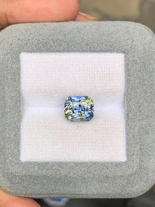 Un saphir bicolore de 5,01 carats signé Bonnot Paris, aux reflets bleu-jaune, est niché dans un écrin gris doux. Des doigts délicatement saisis mettent en valeur les facettes complexes de la pierre, évoquant les éclatantes pierres précieuses du Sri Lanka.