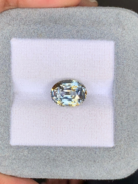 Une main berce délicatement un saphir bicolore de 5,01 carats, légèrement jaune, de Madagascar, niché dans un écrin carré en velours gris. Ce joyau signé Bonnot Paris révèle brillance et clarté, ses facettes captant la lumière.