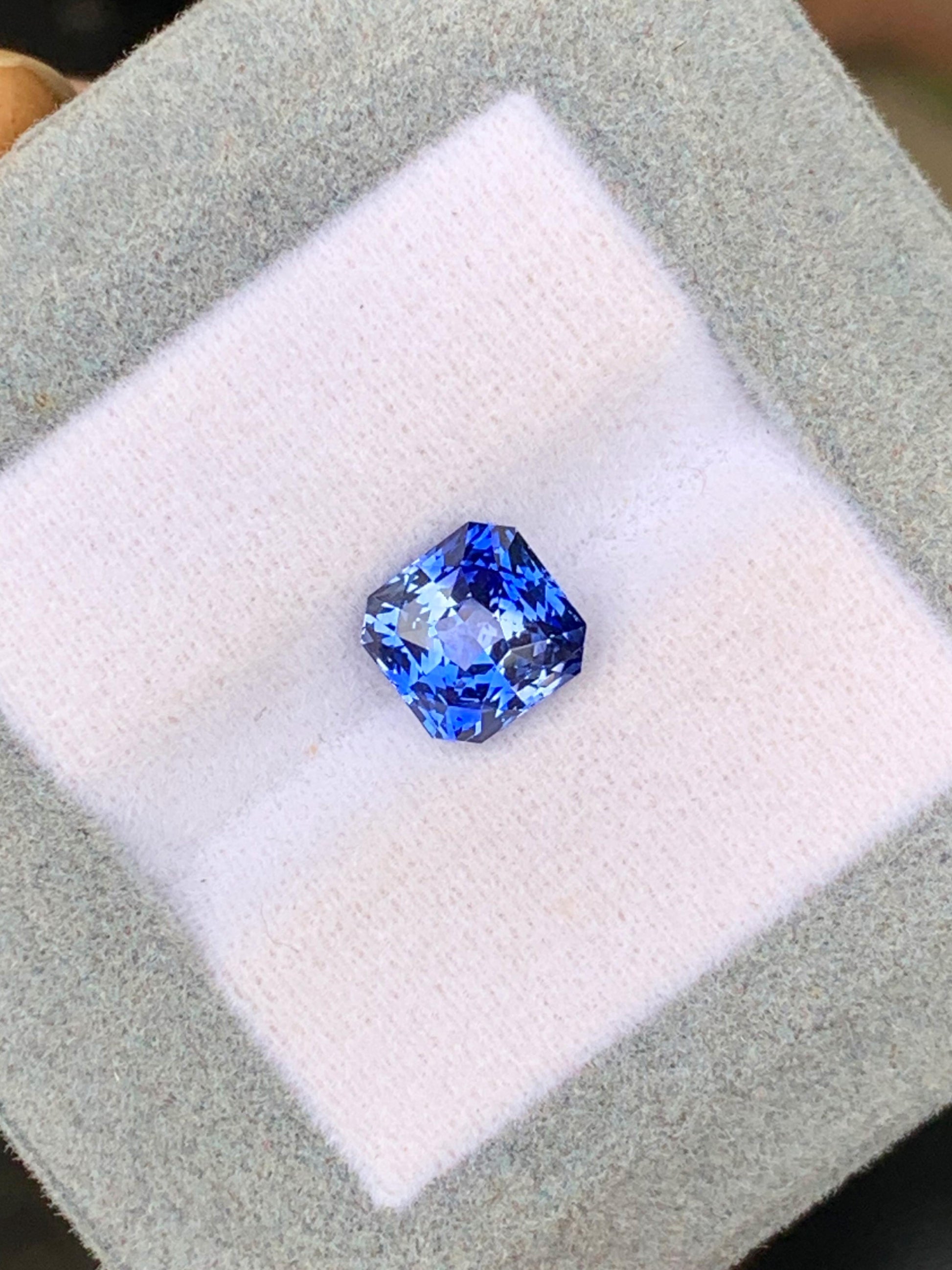 Le Saphir bicolore • 3,02ct de [NOUVEAU] Bonnot Paris, une pierre précieuse carrée d'un bleu vibrant provenant du Sri Lanka, repose sur une surface coussinée gris clair et blanche.