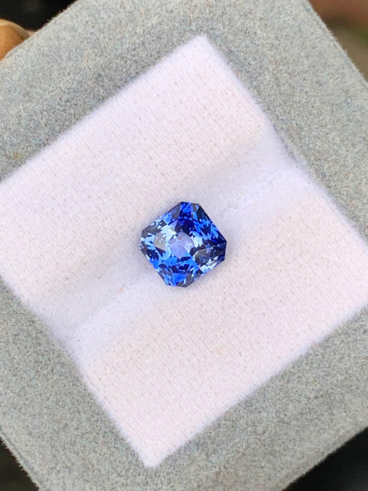 Le Saphir bicolore • 3,02ct de [NOUVEAU] Bonnot Paris, une pierre précieuse carrée d'un bleu vibrant provenant du Sri Lanka, repose sur une surface coussinée gris clair et blanche.