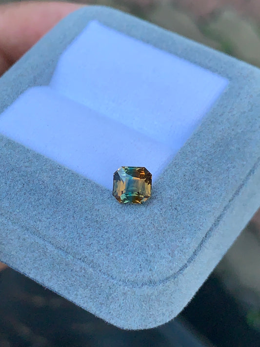 Une main tient un luxueux coffret à bijoux en velours bleu pâle signé Bonnot Paris, exposant le Saphir bicolore de 1,60 carat. Cette pierre taille émeraude présente des nuances vertes et brunes ; ses facettes captent la lumière, révélant une clarté et une vivacité rappelant une forêt d'un vert intense teinté d'orange chaleureux.