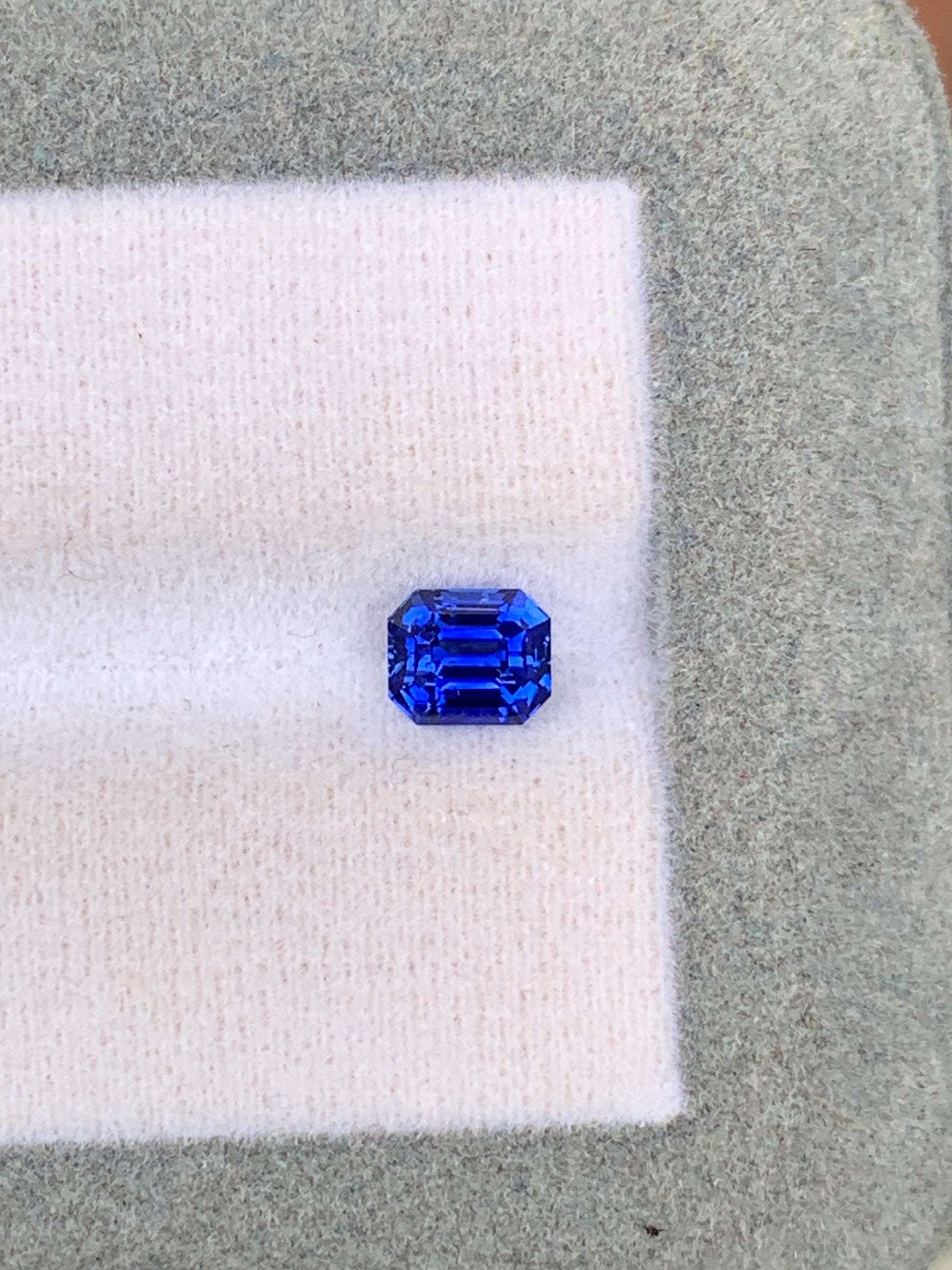 Le Saphir bleu • 1,02ct de [NOUVEAU] Bonnot Paris est une gemme taille émeraude du Sri Lanka, reposant sur un doux tissu blanc et gris, mettant en valeur ses facettes claires et sa riche couleur bleue.