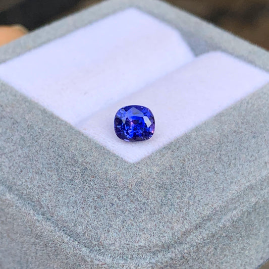 Le Saphir bleu • 1,08ct de Bonnot Paris, une pierre précieuse bleue facettée éclatante du Sri Lanka, est élégamment présenté sur un coussin blanc dans un écrin à bijoux gris clair, incarnant la joaillerie exceptionnelle.