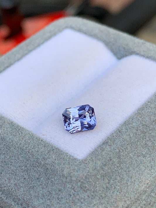 Une pierre bleue rectangulaire facettée de la maison Bonnot Paris, « Saphir bleu • 1,15 ct », est sertie sur un coussin blanc dans un écrin gris ouvert. La pierre reflète la lumière, sublimant sa teinte vibrante, rappelant le charme du Sri Lanka.