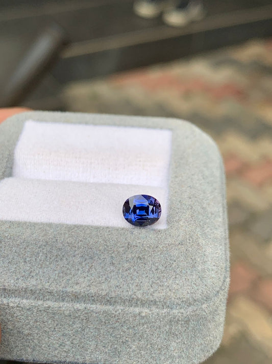 Un saphir bleu de 1,20 carat, taille ovale et éclatant, repose sur un tissu blanc dans un écrin gris ouvert signé Bonnot Paris. Les carreaux texturés et flous en arrière-plan rehaussent l'attrait de ce joyau exquis de Madagascar.