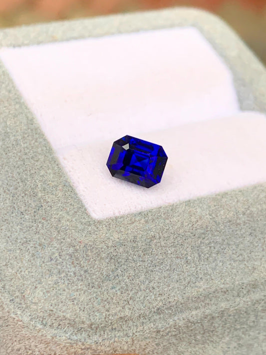 Sur un présentoir à bijoux blanc rembourré, la pierre précieuse Saphir bleu profond taille émeraude rectangulaire • 1,51 ct de [NOUVEAU] Bonnot Paris du Sri Lanka est posée sur un fond flou.