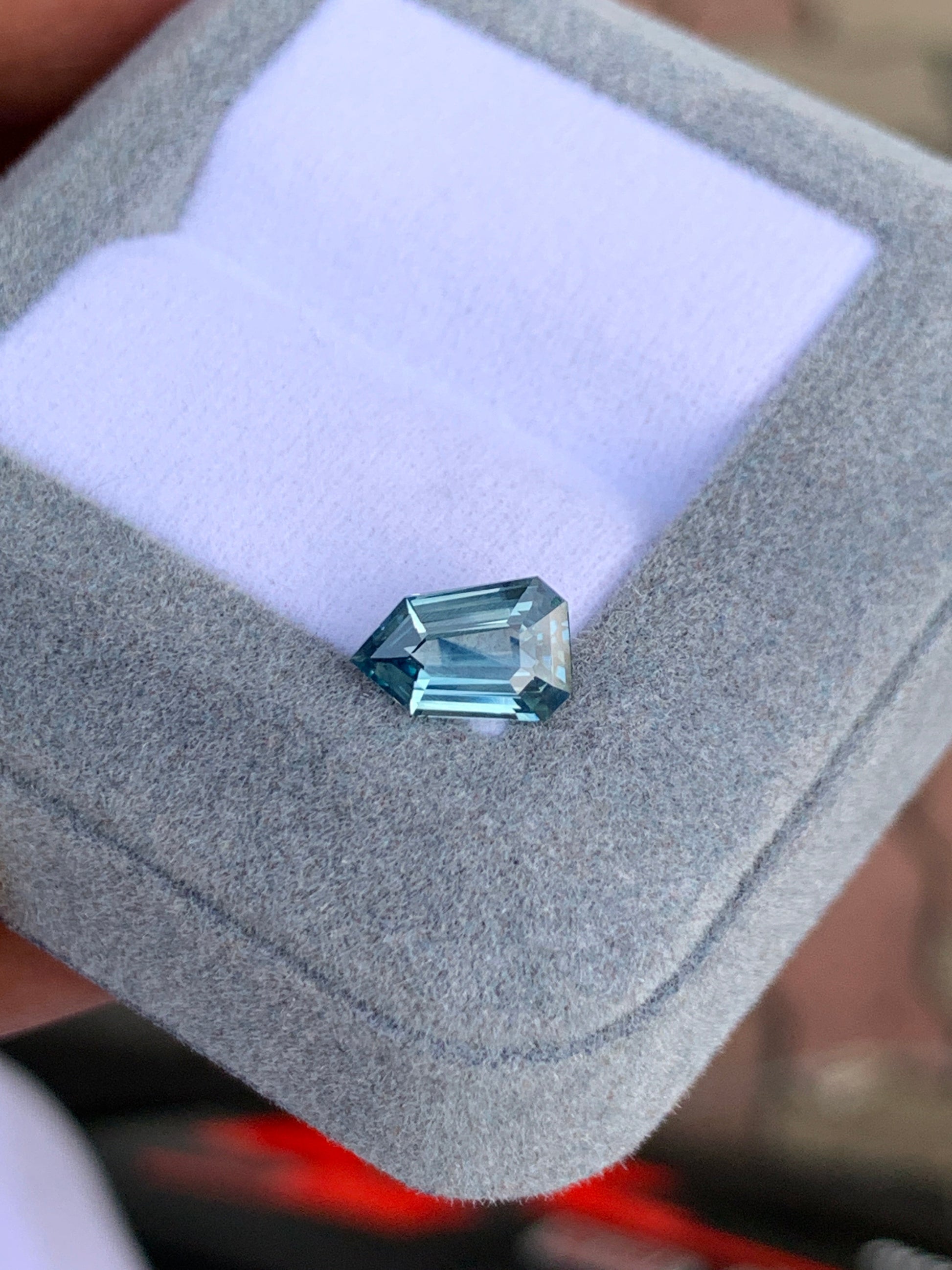Le Saphir bleu Bonnot Paris • 1,59ct, une pierre précieuse bleue étincelante avec une taille géométrique angulaire, est magnifiquement niché dans un coffret à bijoux en velours gris avec un intérieur blanc.
