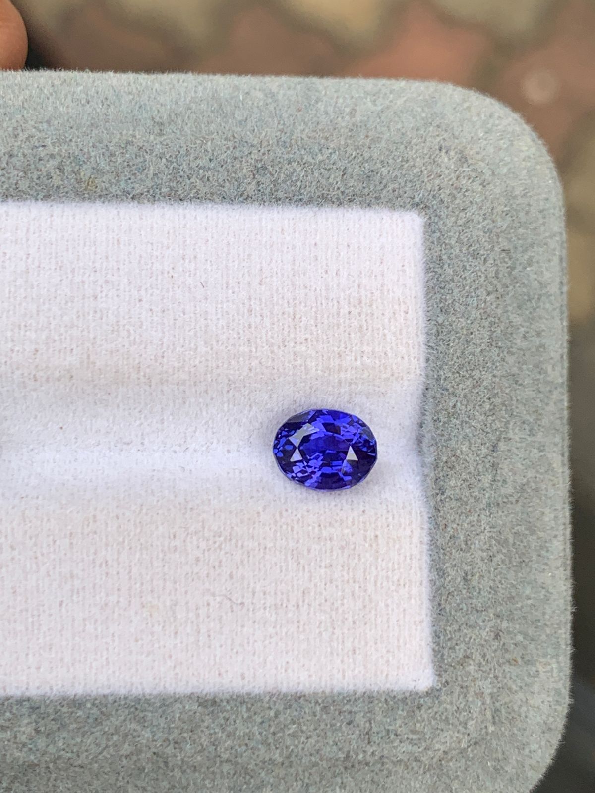 Un Saphir bleu taille ovale de 1,60 ct de [NOUVEAU] Bonnot Paris se trouve dans une boîte gris clair avec un intérieur blanc, ses facettes bleues vibrantes scintillant sur un fond aux tons terreux.
