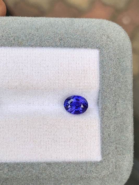 Un Saphir bleu taille ovale de 1,60 ct de [NOUVEAU] Bonnot Paris se trouve dans une boîte gris clair avec un intérieur blanc, ses facettes bleues vibrantes scintillant sur un fond aux tons terreux.