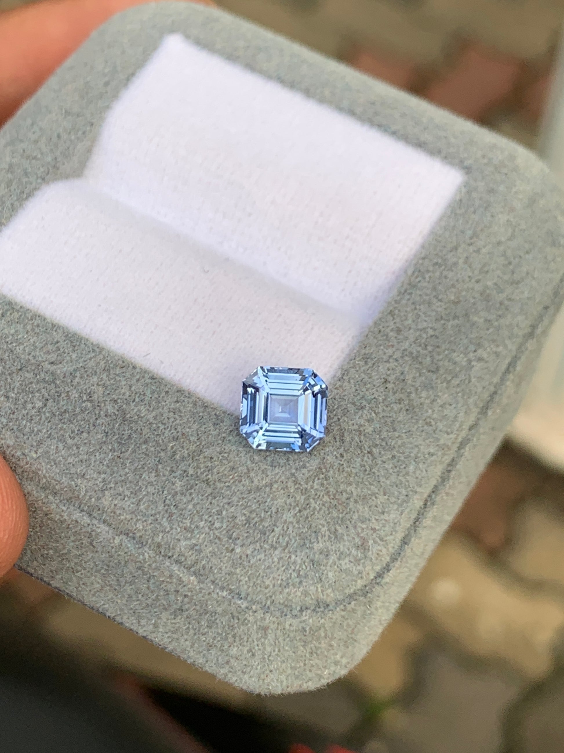 Quelqu'un tient un coffret à bijoux gris contenant un coussin, présentant un Saphir bleu taille carrée facetté de 1,70 carat de Bonnot Paris. L'arrière-plan légèrement flou souligne l'allure sophistiquée de cette joaillerie unique sur le sol en pierre.