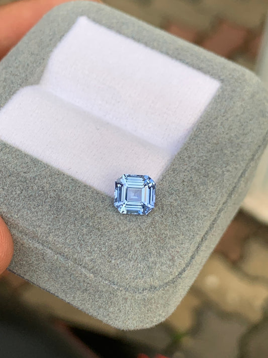 Quelqu'un tient un coffret à bijoux gris contenant un coussin, présentant un Saphir bleu taille carrée facetté de 1,70 carat de Bonnot Paris. L'arrière-plan légèrement flou souligne l'allure sophistiquée de cette joaillerie unique sur le sol en pierre.
