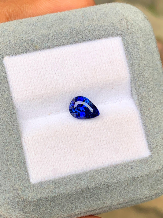 Un saphir bleu poire taille brillant de 1,76 ct de Bonnot Paris, présenté dans un écrin gris clair matelassé, est orné d'une pierre précieuse éclatante, évoquant la pierre d'exception sri-lankaise. Il est soigneusement centré sur un carré de tissu blanc.