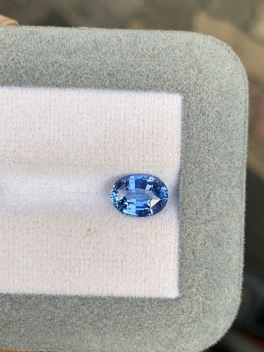 Un saphir bleu étincelant de 2,10 ct de Bonnot Paris en provenance du Sri Lanka repose sur un coussin blanc avec une bordure gris clair, sa surface ovale facettée rehaussant la brillance sur le fond - un chef-d'œuvre de l'artisanat de la joaillerie.