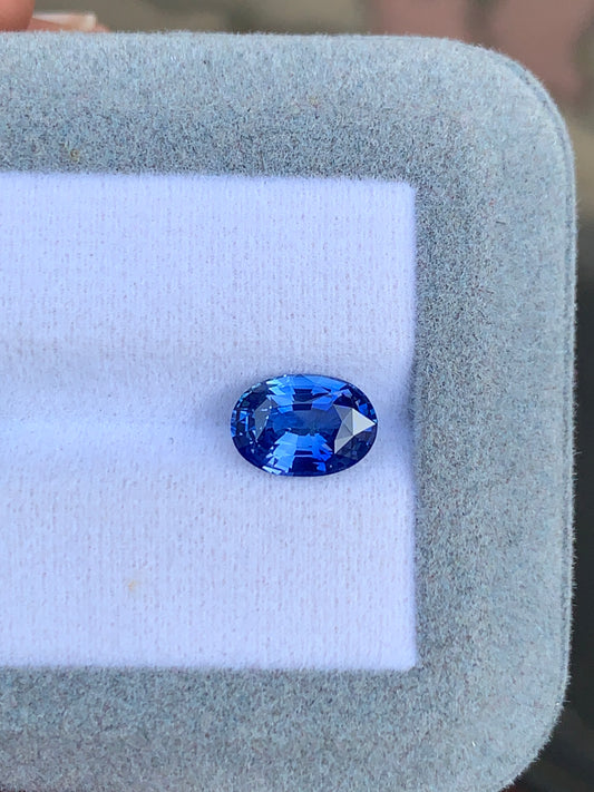 Un Saphir bleu de 2,51 carats de Bonnot Paris brille de mille feux sur une surface coussinée blanche, entourée d'un tissu gris clair, soulignant son allure exquise.