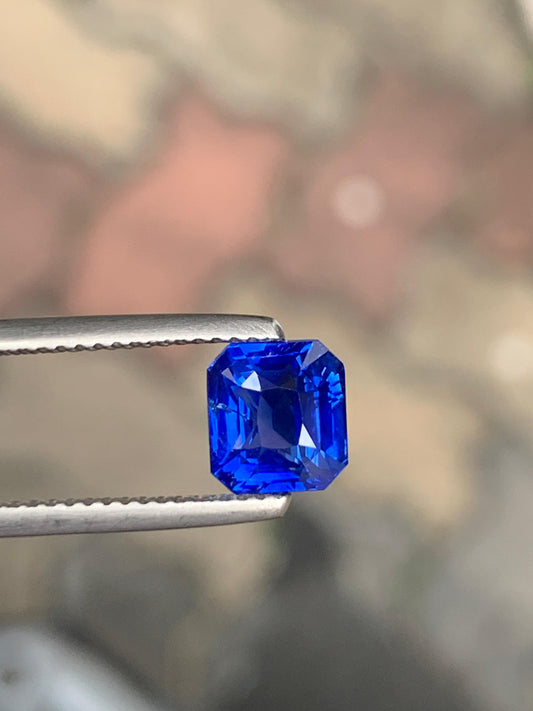 Gros plan sur le Saphir bleu • 2,53 ct de [NOUVEAU] Bonnot Paris, un saphir sri-lankais d'un bleu éclatant, maintenu par une pince métallique. La teinte profonde et non chauffée de cette pierre taille émeraude se détache sur un fond flou, soulignant sa clarté et sa couleur incomparables.